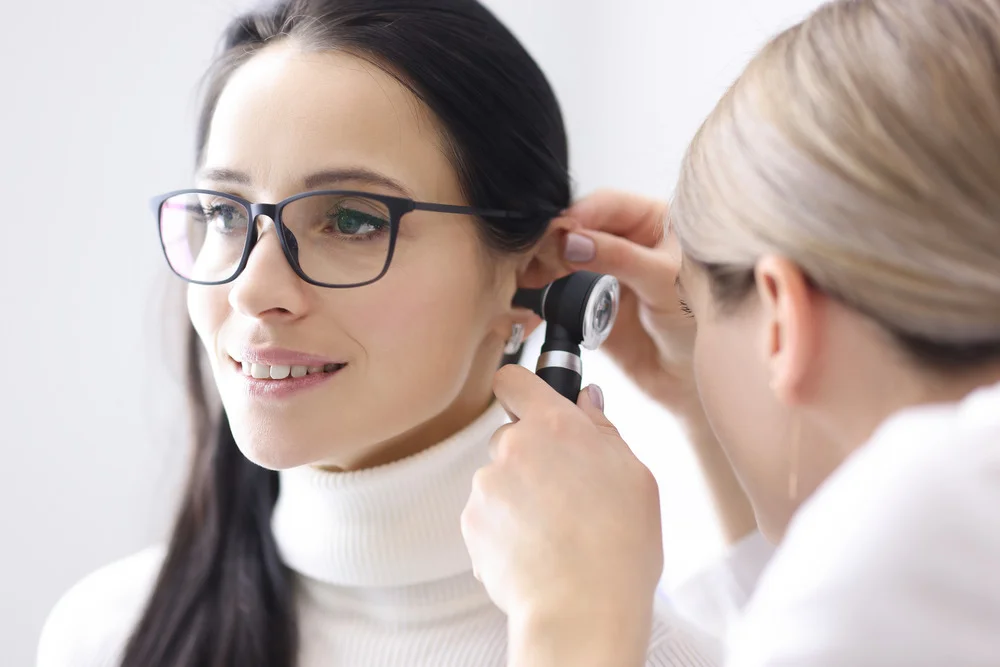 Woman with long brown hair wearing glasses and a white turtleneck having otoscopy performed by healthcare professional/ audiologist